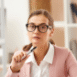 act-2 Woman in glasses holding a pen, contemplating, in a professional office setting, reflecting Solo Performance LLC's focus on leadership and training in the automotive sales industry.