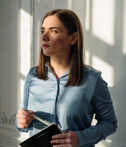 Woman in a blue blouse holding a notebook, reflecting on dealership performance and training strategies, with light streaming through a window.
