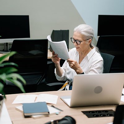 Older woman reading documents at a desk with a laptop, surrounded by notebooks and a plant, representing a collaborative discussion in a professional setting for IT services and business transformation.
