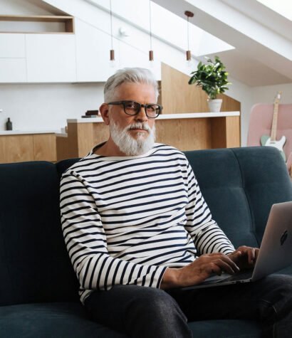 Older man with glasses and a striped shirt working on a laptop in a modern living space, reflecting a professional environment related to training and consulting services.