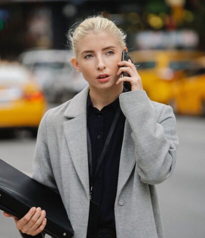 Professional woman in gray coat on phone, holding a binder, with yellow taxis in the background, representing business and communication in urban settings.