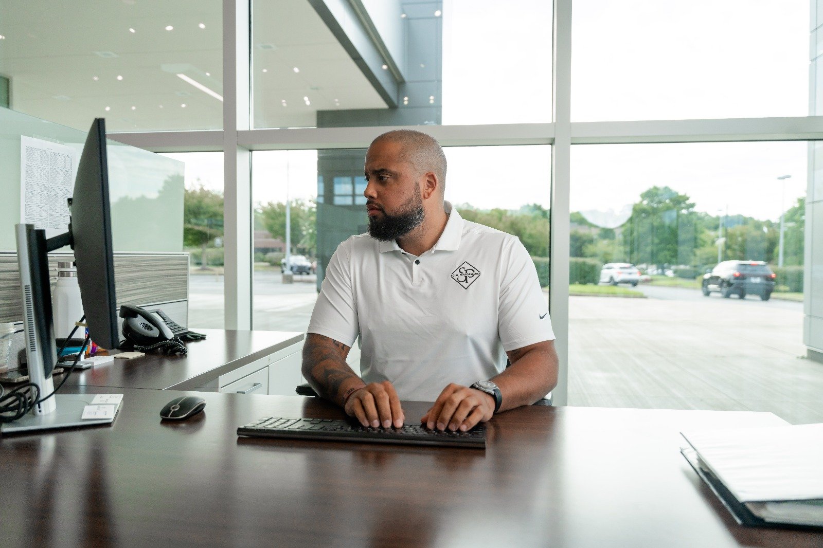 Man in a white polo shirt at a desk using a computer, focused on automotive sales training tasks in a modern dealership environment.