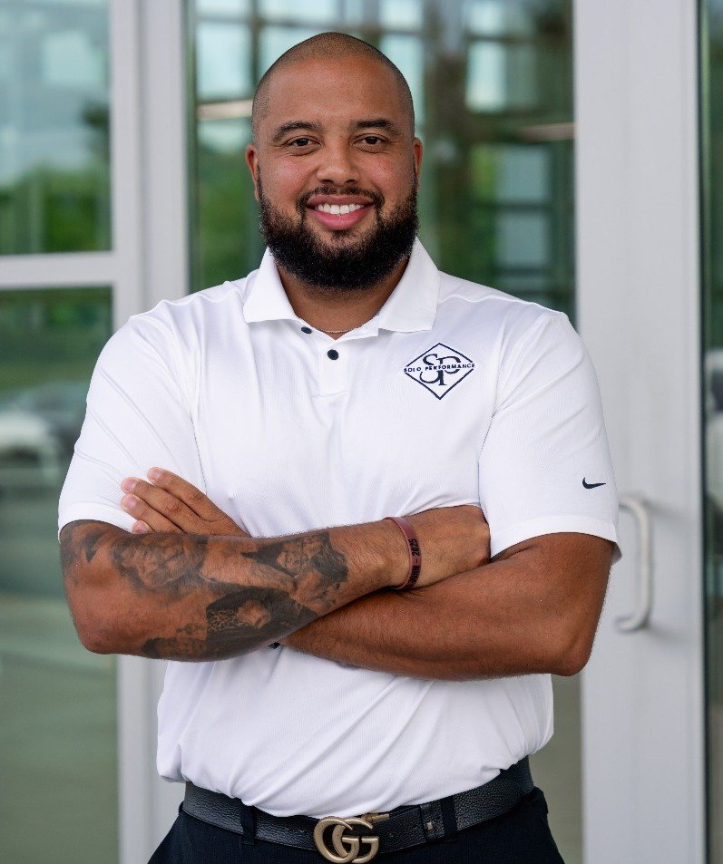 Man smiling with arms crossed, wearing a white polo shirt with Solo Performance LLC logo, standing in front of glass doors, representing training and consulting services for auto dealerships.