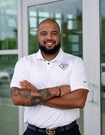 Frederick Edmonson, founder and CEO of Solo Performance LLC, smiling with arms crossed, wearing a white polo shirt with a logo, standing in front of a dealership setting, representing automotive sales and finance training expertise.
