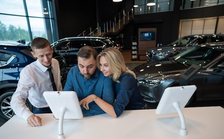 Three dealership employees engaged in a discussion around a tablet, surrounded by cars in a modern showroom, illustrating collaboration and customer interaction in automotive sales training.
