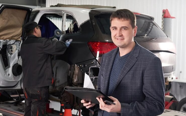 Man in a blazer holding a clipboard, standing in an automotive workshop with a mechanic working on a vehicle, illustrating automotive sales management and training context.