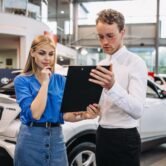 Woman and man discussing automotive sales strategies while reviewing a document in a car dealership setting.
